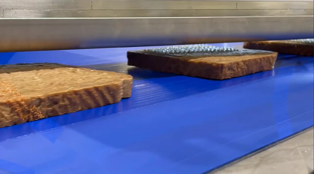 Slices of brown bread on a blue conveyor under the machine, the middle slice with rows of small butter drops on top