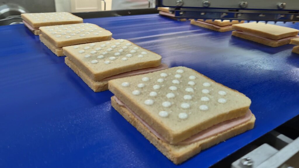 Sandwiches on a blue conveyor behind the depositing head, each top slice with a grid of white sauce drops